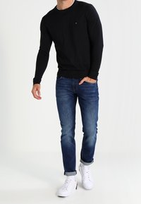 Hombre con suéter negro de manga larga, jeans azules con puños, zapatillas blancas, de pie con una mano en el bolsillo contra un fondo blanco.
