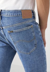 Los jeans de mezclilla azul cuentan con una tela texturizada, un parche trasero de cuero marrón y bolsillos traseros de doble costura con un diseño curvado.