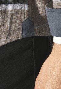 Karierte Hemd in Braun und Grau mit einem einfarbigen dunkelgrauen Kragen und einem auffälligen Patch-Detail am Saum, kombiniert mit schwarzen Hosen.