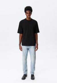 Mann iført svart oversized T-skjorte, lyseblå jeans og svarte støvler, står mot en hvit bakgrunn.