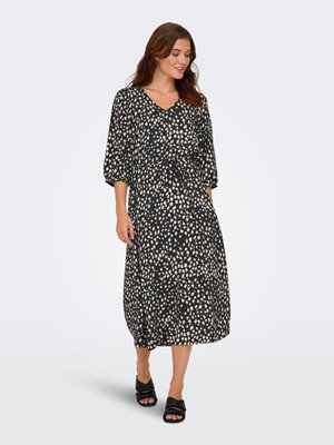 Femme portant une robe midi noire avec des taches blanches irrégulières, manches trois-quarts, taille ceinturée et talons ouverts noirs, debout et souriante.
