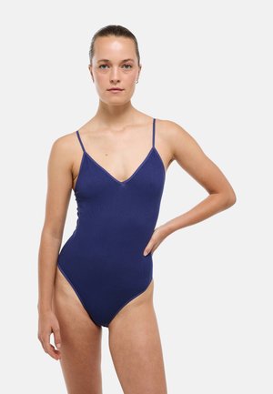 Femme aux cheveux attachés porte un body sans manches bleu marine uni avec de fines bretelles, debout avec une main sur la hanche.