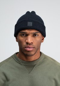 Donkerblauwe gebreide beanie met een geribbelde textuur en een klein zwart logo label. Draag het met een groene sweater.