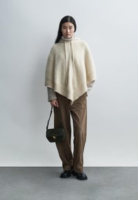 Kremni pončo iz mehkega volnenega blaga z teksturirano površino, kombiniran z lahkotnim puloverjem, rjavimi žametnimi hlačami in temno zeleno torbico za ramo.