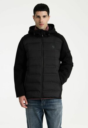 Mann trägt schwarze gesteppte Kapuzenjacke und blaue Jeans und steht vor weißem Hintergrund.