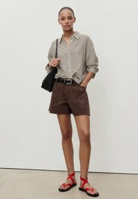 Shorts marron et chemise à boutons crème rayée. Ceinture et sac noirs. Sandales rouges à lanières. Textures lisses, coupe décontractée. Fond minimaliste.