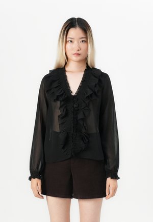 Svart chiffonbluse med volangdetaljer ved halsen og mansjettene, semi-transparent stoff, lange ermer og knappelukking foran. Kombinert med mørke shorts.