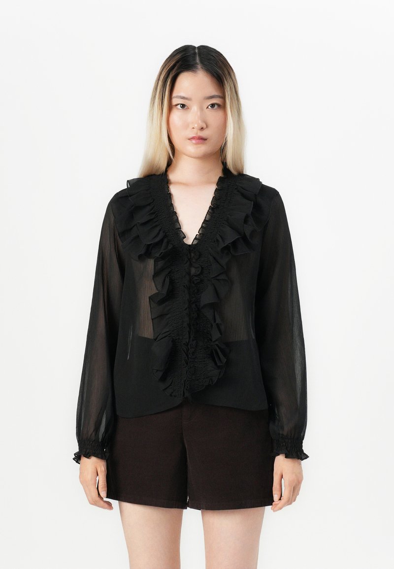 Svart chiffonbluse med volangdetaljer ved halsen og mansjettene, semi-transparent stoff, lange ermer og knappelukking foran. Kombinert med mørke shorts.