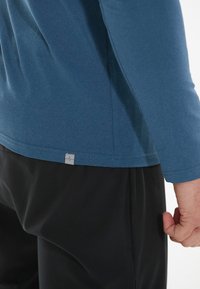 Long-sleeve, blue athletic shirt in a soft, breathable fabric with a seam detail and a gray logo tag at the hem, paired with black pants.