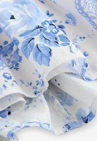 Balta audinys su mėlynu gėlių raštu, vaizduojantis lapus ir gėles, matomos audinio kraštinės ir siuvimo detalės.