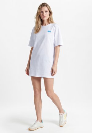 Jeune femme portant une robe T-shirt blanche oversize avec le texte « American Vintage », debout avec une jambe légèrement avancée, baskets blanches.