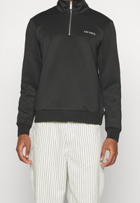 Sudadera negra con cremallera y cuello alto, que presenta un panel en contraste y el logo "LES DEUX" en el pecho, hecha de un tejido suave. Pantalones blancos a rayas.