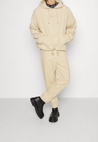 Felpa in corduroy beige con coulisse e tasca frontale, abbinata a pantaloni tapered coordinati. Stivali neri alla caviglia con suole testurizzate.