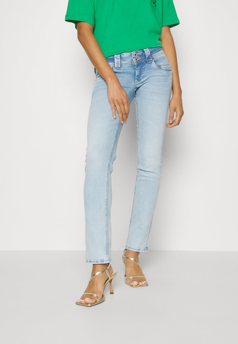 Helle blaue Jeans aus Denim mit schlanker Passform, fünf Taschen und einer leichten Waschung. Kombiniert mit einem grünen Oberteil und goldenen Absatzsandalen.