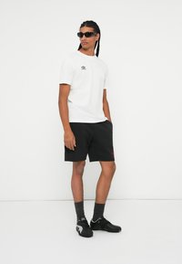 Camiseta blanca de manga corta con logo, combinada con pantalones cortos negros que tienen un logo rojo; los calcetines grises y las zapatillas deportivas negras completan el look.