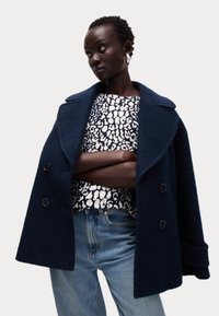 Námořnický vlněný kabát s velkými klopami a dvojřadovým zapínáním, přehozený přes bílý a černý vzorovaný top a modré džíny.