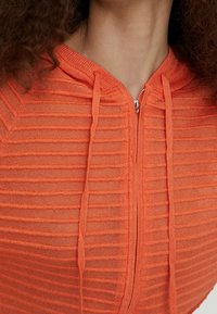 Orange zip-up hoodie with vertical ribbed texture, featuring drawstrings and a metallic zipper. Lightweight, fitted design with a hood.