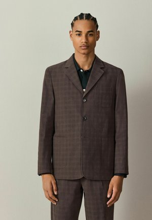 Young man with braided hair wearing a dark brown checked suit jacket and matching trousers, standing against a plain light wall.
