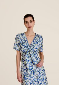 Blouse à motifs bleu et blanc avec un devant à nouer, des manches courtes et un décolleté en V profond. Présente des formes abstraites et une texture lisse.