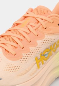 Chaussure de sport légère couleur pêche avec un dessus en mesh, un système de laçage et un logo jaune. Présente une semelle sculptée et des accents subtils de bleu dans le design.