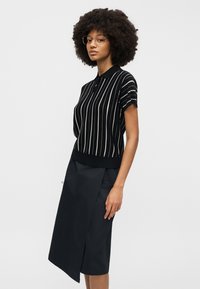 Femeie cu păr creț, purtând ochelari, un top negru cu mâneci scurte și guler, cu dungi verticale albe, și o fustă neagră asimetrică, până la genunchi.