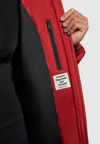 Rode gewatteerde jas met een zwart voering; beschikt over een ritszak en een label met de tekst: "Een warme omhelzing gemaakt van stof."
