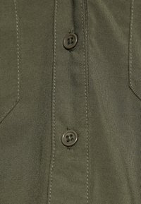 Olivgrüner Hemdenstoff mit glatter Textur, ausgestattet mit zwei schwarzen Knöpfen und sichtbaren Nähten entlang der Nähte und Taschen.