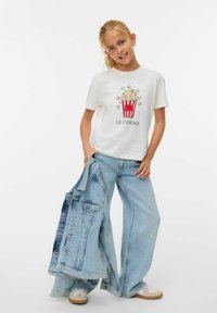 T-shirt blanc à manches courtes présentant un graphique de seau de popcorn rouge et blanc avec le texte "LE CINÉMA" ; associé à un jean en denim bleu clair à jambes larges.
