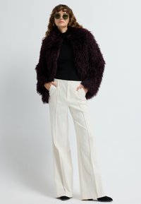 Kvinde iført en mørk lilla, fuzzy jakke, sort turtleneck, hvid højtaljeret bukser med brede ben, sorte sko, solbriller og guldbelagte hoop øreringe.