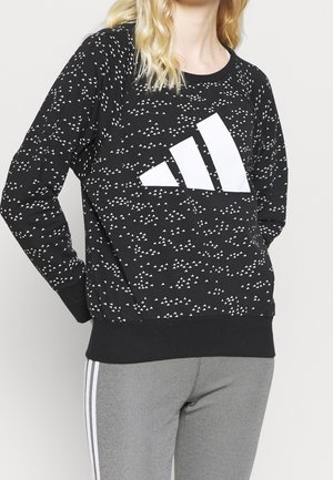 Osoba nosząca czarną bluzę z białymi, rozproszonymi trójkątnymi wzorami oraz dużym białym logo Adidas, w połączeniu z szarymi spodniami z białymi bocznymi paskami.