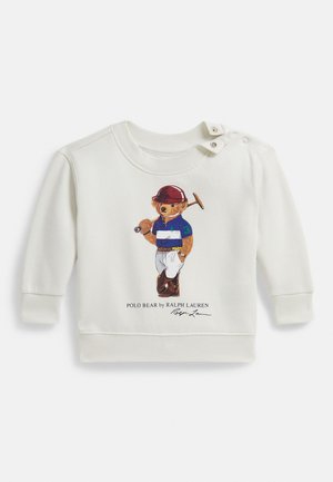 Kremfarget genser laget av mykt stoff, med en fargerik illustrasjon av en bjørn i en pikétrøye. Designet med trykknapper på skulderen.