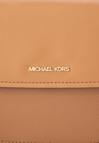 Geantă de mână din piele maro deschis, cu textură netedă, logo "MICHAEL KORS" într-o nuanță aurie și cusături precise. Design compact și dreptunghiular.