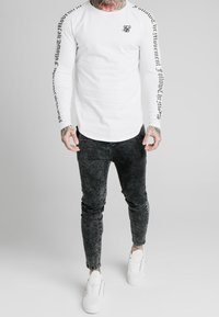 Vit långärmad skjorta med svart text på ärmarna, kombinerad med svarta smala jeans och vita sneakers. Diskret logotyp på bröstet.