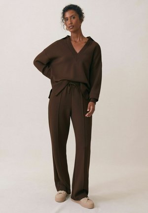 Kobieta o kręconych włosach w ciemnobrązowym swetrze z dekoltem w serek i dopasowanych szerokich spodniach, nosząca beżowe trampki, stojąca na jednolitym tle.