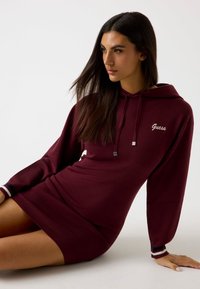 Vestito con cappuccio color borgogna a maniche lunghe, realizzato con un tessuto morbido. Presenta il logo "Guess" sul petto e dettagli a righe sui polsini delle maniche.
