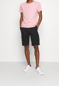 Camiseta de algodón rosa claro con cuello redondo y un pequeño logo, combinada con pantalones cortos cargo negros con bolsillos laterales. Usada con zapatillas blancas.