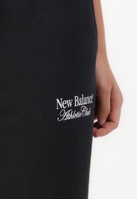 Melnas sporta bikses no mīksta auduma, ar baltu logotipa drukājumu "New Balance Athletic Club" sānos.
