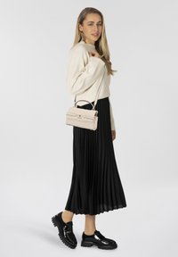 Femme avec de longs cheveux blonds portant un pull crème, une jupe midi plissée noire, des chaussures à plateforme noires brillantes et tenant un sac à épaule matelassé beige.