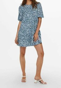 Šaty s květinovým vzorem, krátkými objemnými rukávy, modro-bílé barvy, volný střih, nad kolena, v kombinaci s bílými sandály na blokovém podpatku.