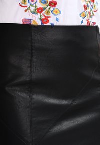 Černá sukně z umělé kůže s hladkou texturou, geometrickými švy a přiléhavým střihem, v kombinaci s bílou vyšívanou blůzou.