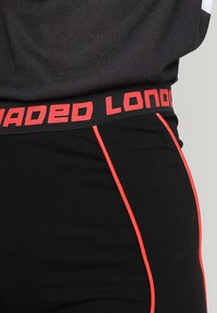 Černé legíny s červeným lemováním a červeným pasem s nápisem "LOADED LONDON". Vyrobené z pružného, hladkého materiálu pro komfort a pohodlné nošení.