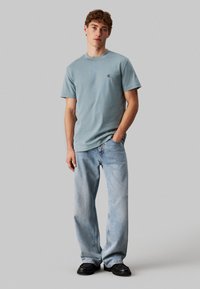 Camiseta de algodón azul claro con mangas cortas, que presenta un pequeño logotipo en el pecho. Combinada con jeans de mezclilla de lavado claro y zapatos negros.