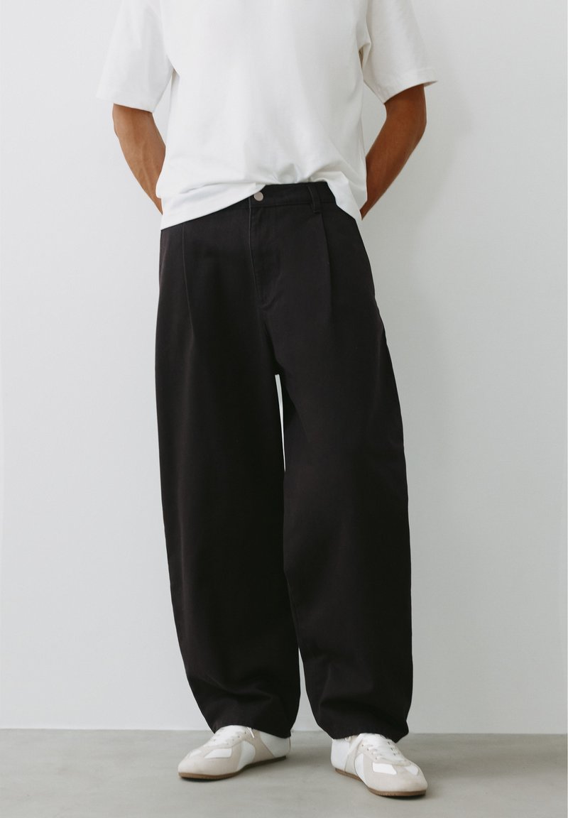 Personne portant un pantalon noir ample, une chemise blanche à manches courtes et des baskets blanches, debout devant un fond blanc uni.