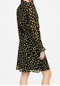 Langärmliges Kleid aus transparentem schwarzem Material mit gelben Polka-Dots. Es verfügt über eine geraffte Taille und gerüschte Ärmelabschlüsse.