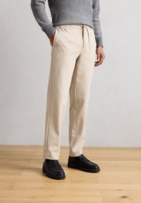 Pantaloni sportivi beige con vita elastica e coulisse, dotati di tasche laterali e vestibilità rilassata. Abbinati a scarpe slip-on nere.