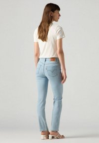 Jeans de mezclilla en azul claro con un corte ajustado, con un diseño clásico de bolsillo trasero y una etiqueta de cuero marrón. Combinados con una camiseta blanca y sandalias.