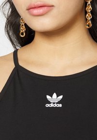 Canotta nera con scollatura a coste e logo Adidas bianco. Visibili orecchini a catena dorati, che aggiungono un contrasto metallico.