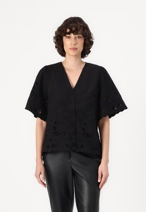 Blouse noire brodée avec un col en V, des manches courtes bouffantes et des motifs floraux découpés complexes. Associée à un pantalon en cuir noir.