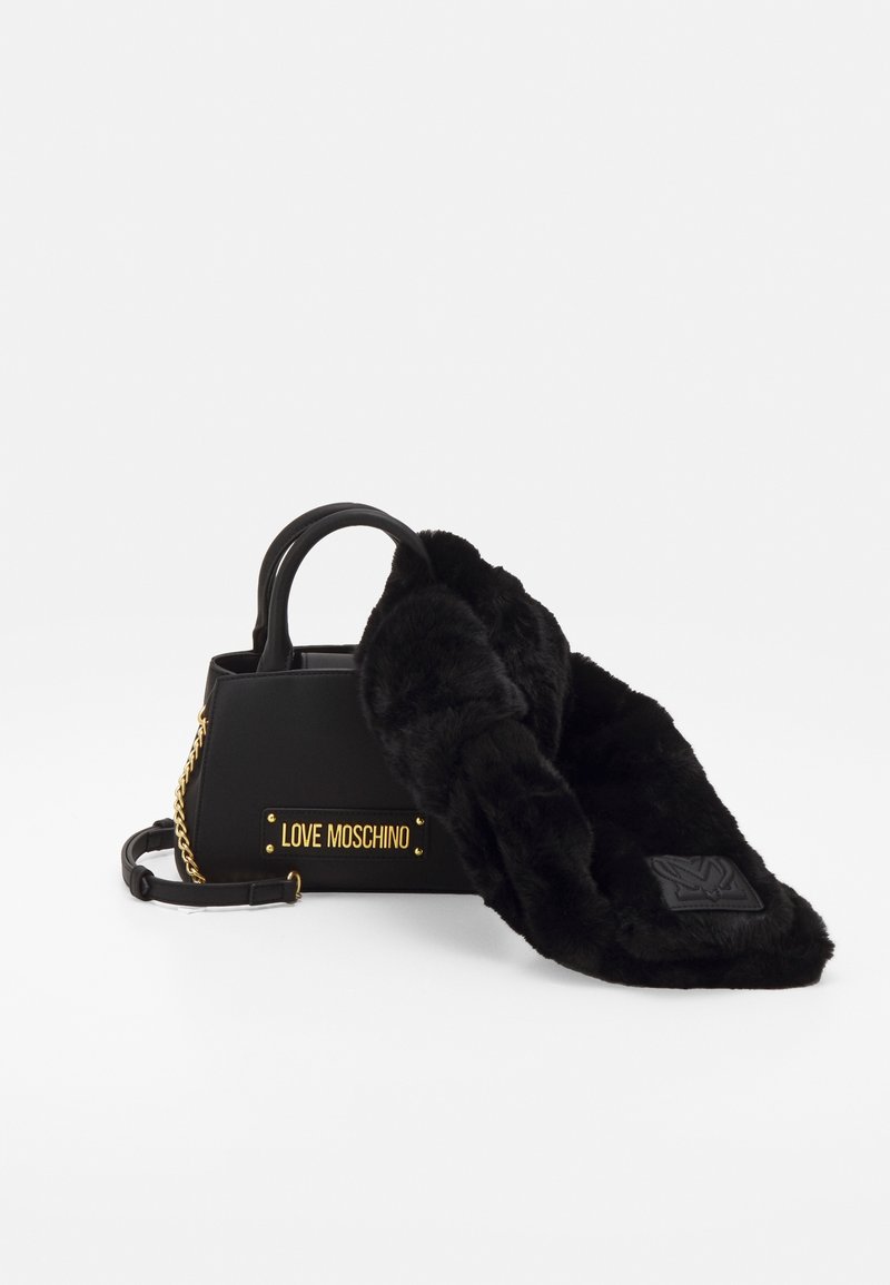 Musta Love Moschino -käsilaukku, jossa on kultaketju, mustat kahvat ja pitkä musta keinoturkishuivi, joka on aseteltu sen päälle valkoisella taustalla.