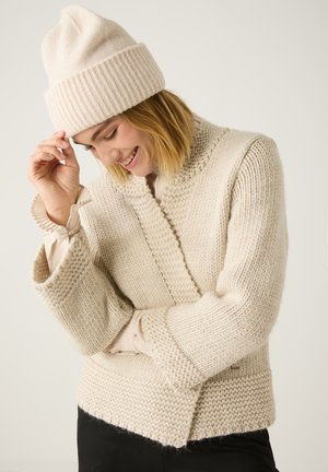 Beige gestrickter Pullover mit weiten Ärmeln und strukturiertem Muster, kombiniert mit einer hellbeigen gerippten Mütze, vor einem schlichten Hintergrund.